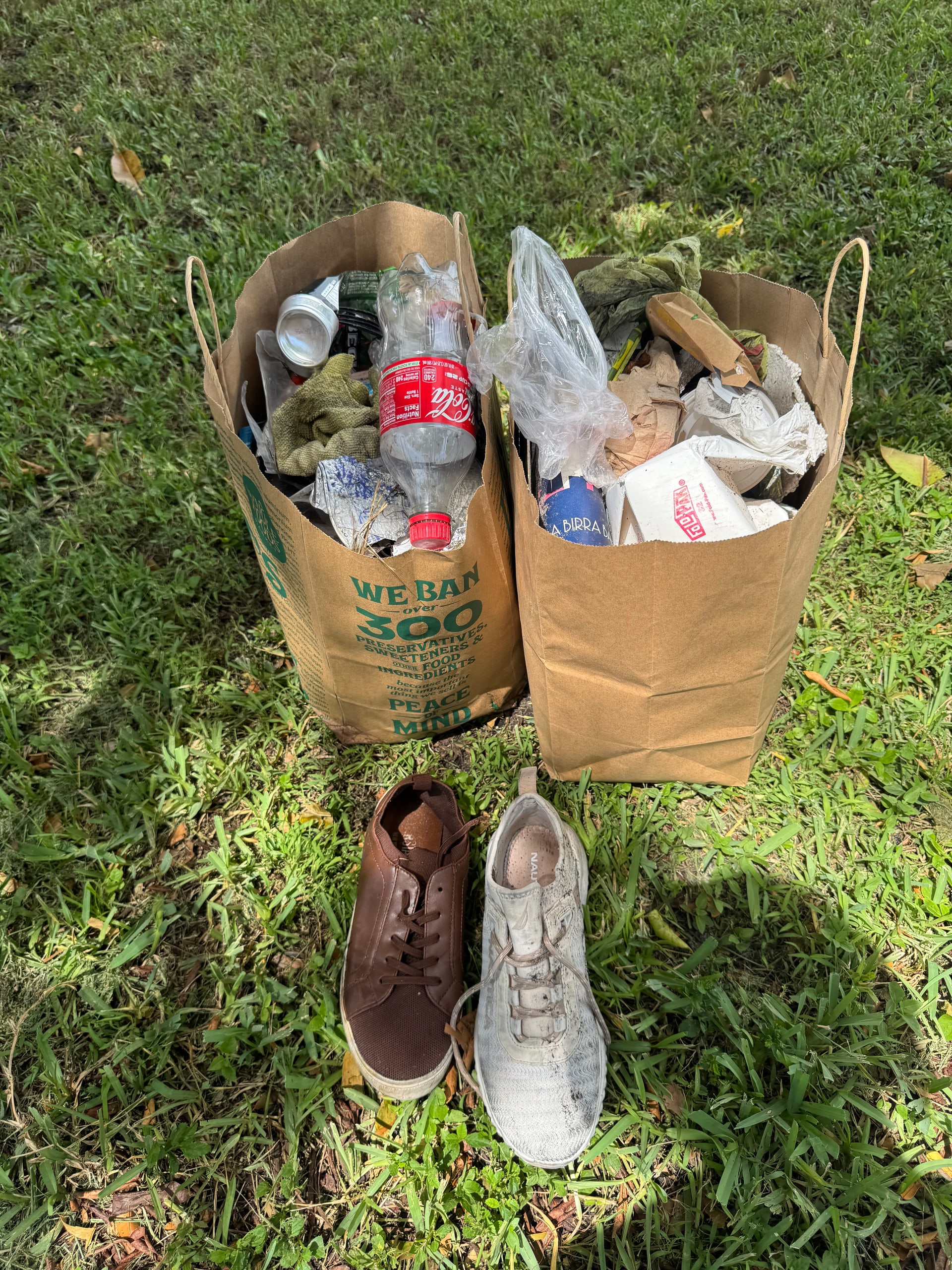 Two bags of bottles, cans, and packaging collected on the grass next to two discarded shoes